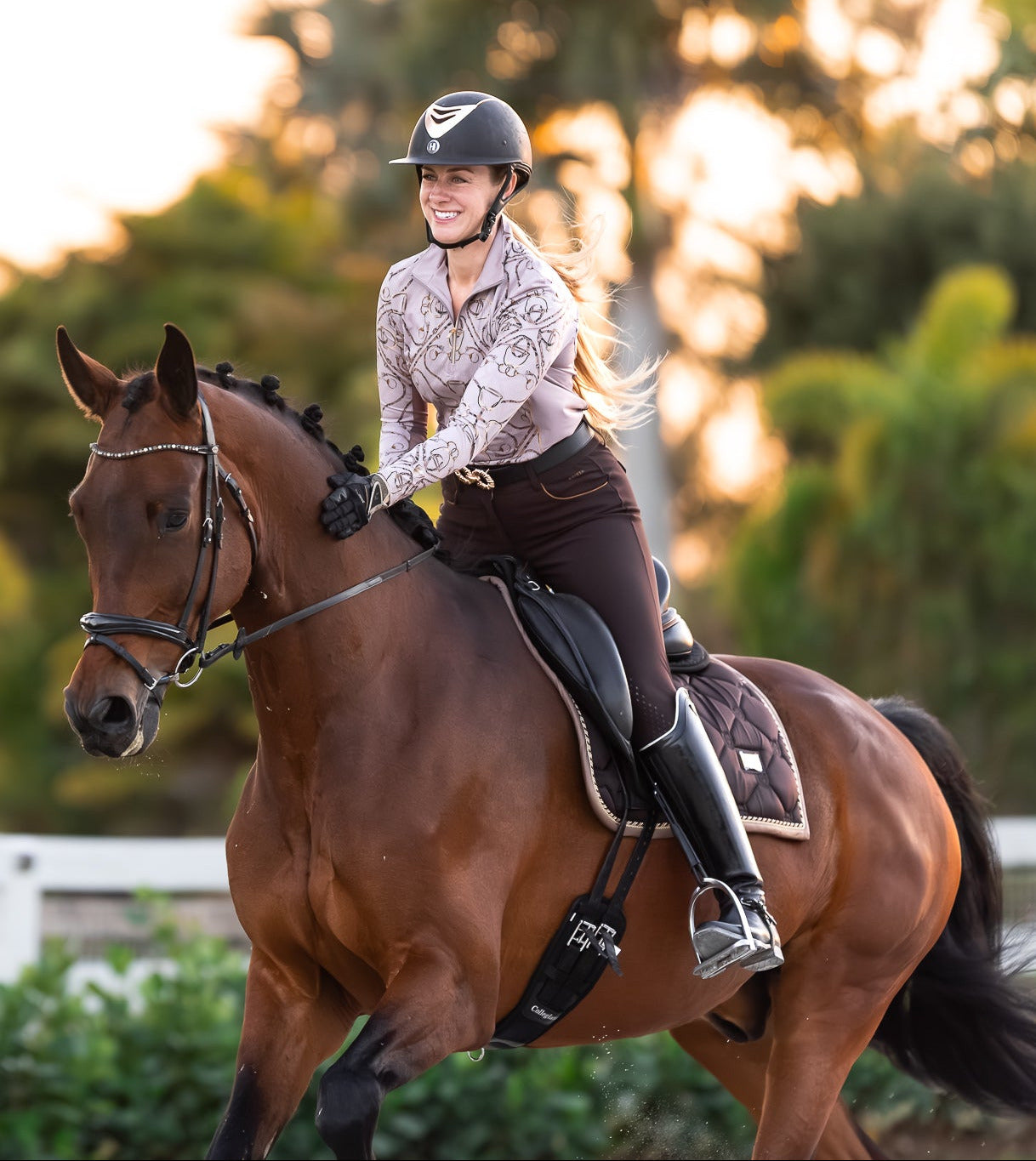 Woman riding a brown horse in an outdoor setting with trees and a white fence in the background.