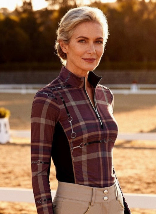 Woman in equestrian attire standing in an outdoor arena with a horse in the background.