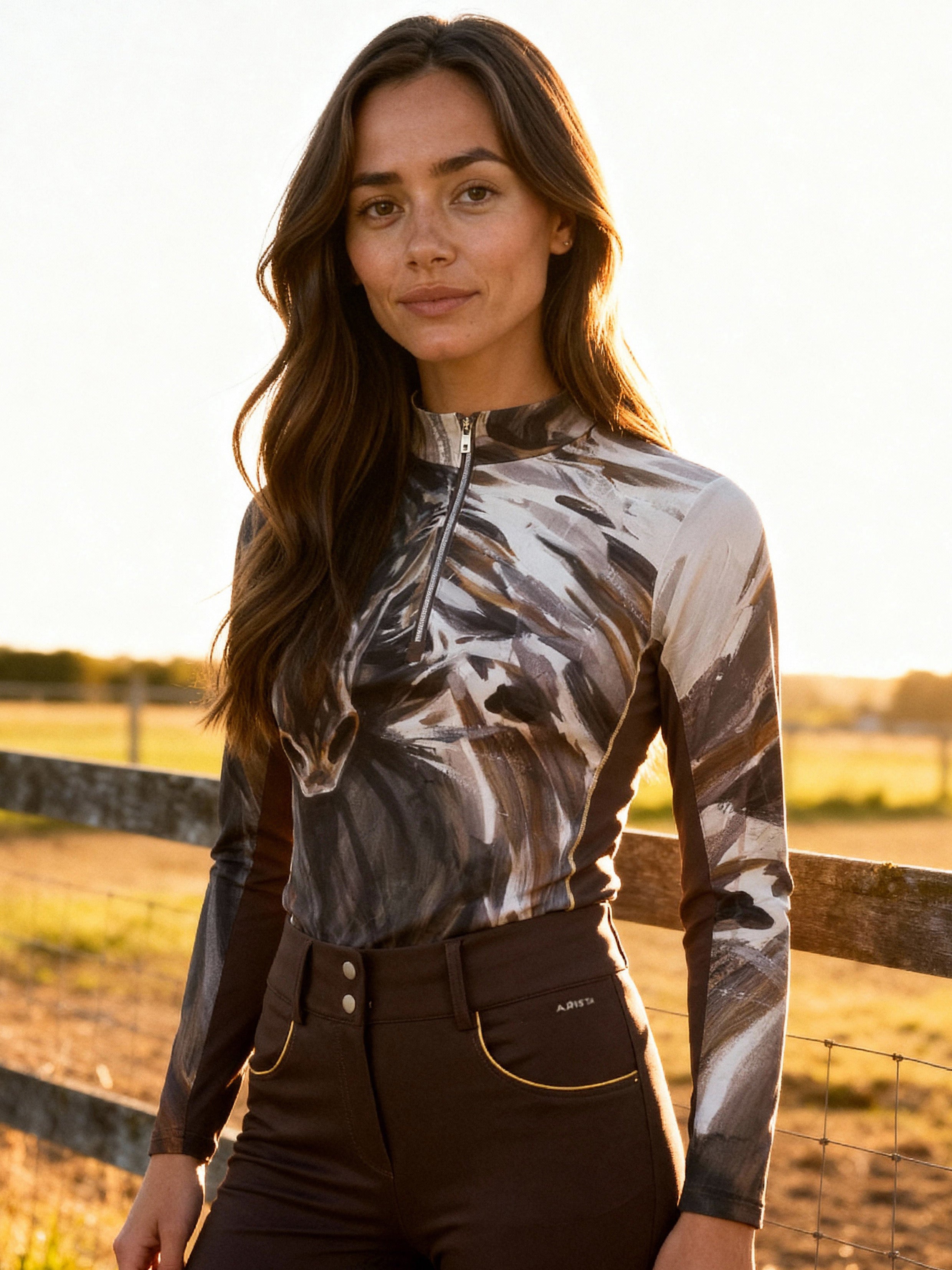 Woman wearing a horse painting long-sleeve shirt and dark pants in a field with a wooden fence.