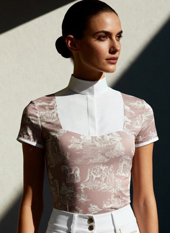Woman wearing a mauve tolie top and white pants against a light wall.