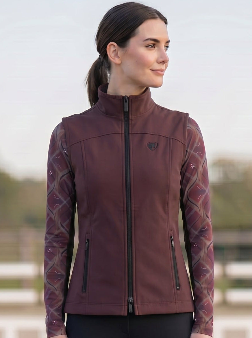 Woman in equestrian attire standing on a gravel surface with equestrian equipment in the background.