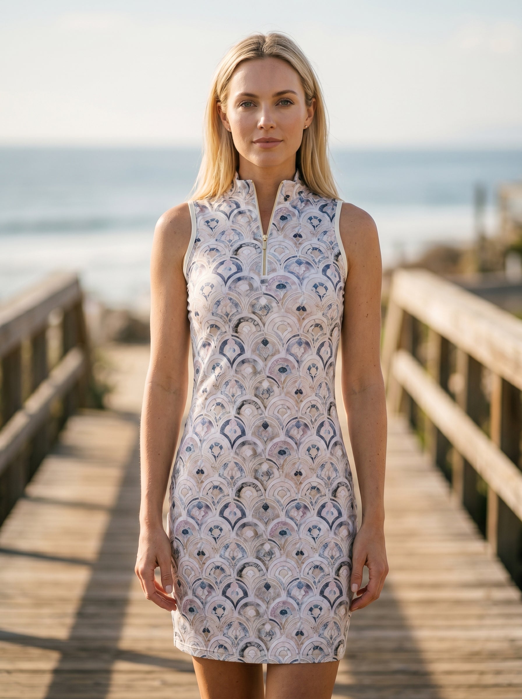 Woman wearing a sleeveless dress with a pattern on a wooden boardwalk by the ocean.