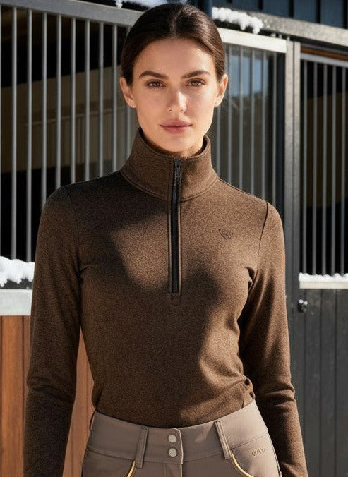 Woman in equestrian attire standing in front of horse stables with snow on the ground