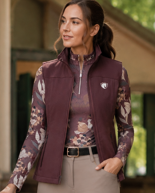 A woman in equestrian attire walks outdoors beside a horse on a stone path. She wears a burgundy vest over a floral long-sleeve shirt, beige riding breeches, and black knee-high boots, with a stable and greenery visible in the background.