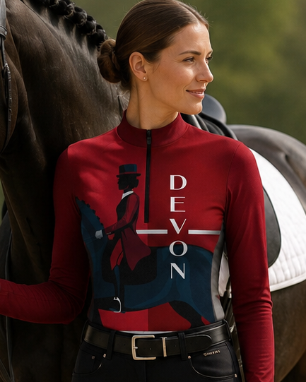 A woman in equestrian apparel stands beside a dark horse in an outdoor riding arena. She wears a burgundy long-sleeve top with a large navy and white graphic and the word 'DEVON' printed vertically, paired with black breeches and tall black riding boots.