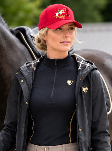 A woman in equestrian attire, including a red cap, black jacket, black top, beige breeches, and black riding boots, walks alongside a black horse on a gravel path in an outdoor stable setting.