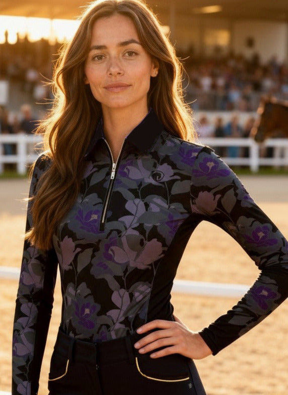 Woman in equestrian attire standing in an outdoor arena with horses in the background