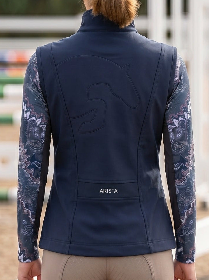 Person wearing equestrian attire with a navy vest and beige pants in an outdoor arena.
