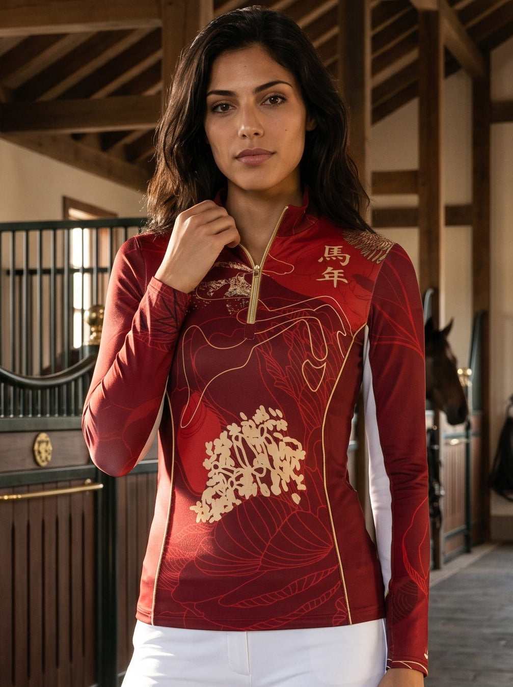 A woman stands in an indoor horse stable wearing a red long-sleeve equestrian-style top with gold embroidery and white riding pants with black boots.
